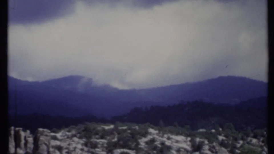 1971 California 8mm view of hazy blue mountain layers, overcast sky, rocky outcrops, and scrub vegetation in the foreground.