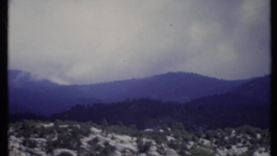 1971 California home-movie frame with low cloud cover, dark mountain ridges, rocky ground, and scattered shrubs.