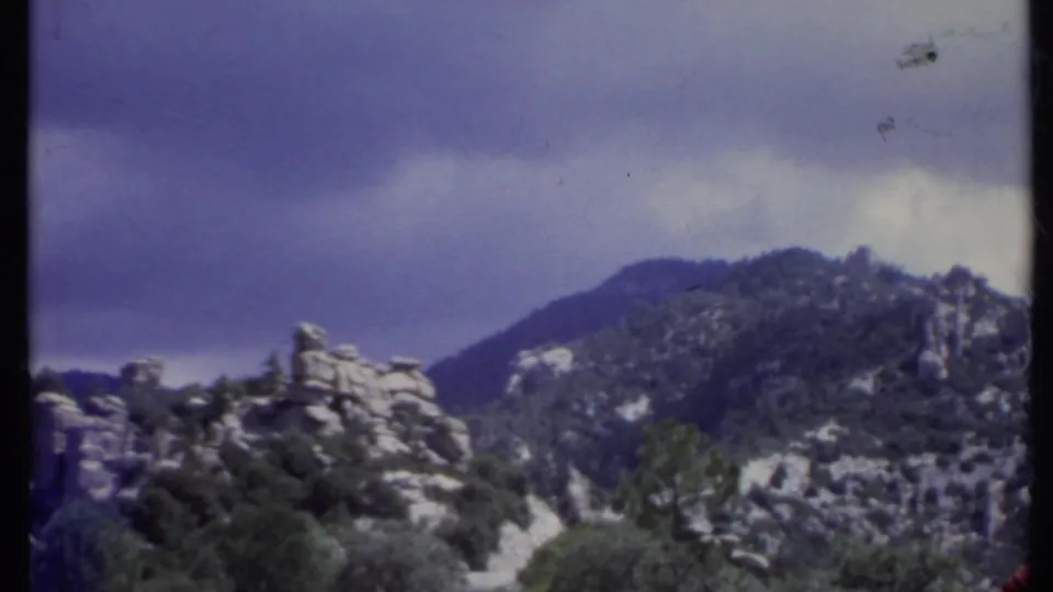 1971 California 8mm frame showing jagged rock formations, a distant mountain slope, shrubs, and a dramatic overcast sky.
