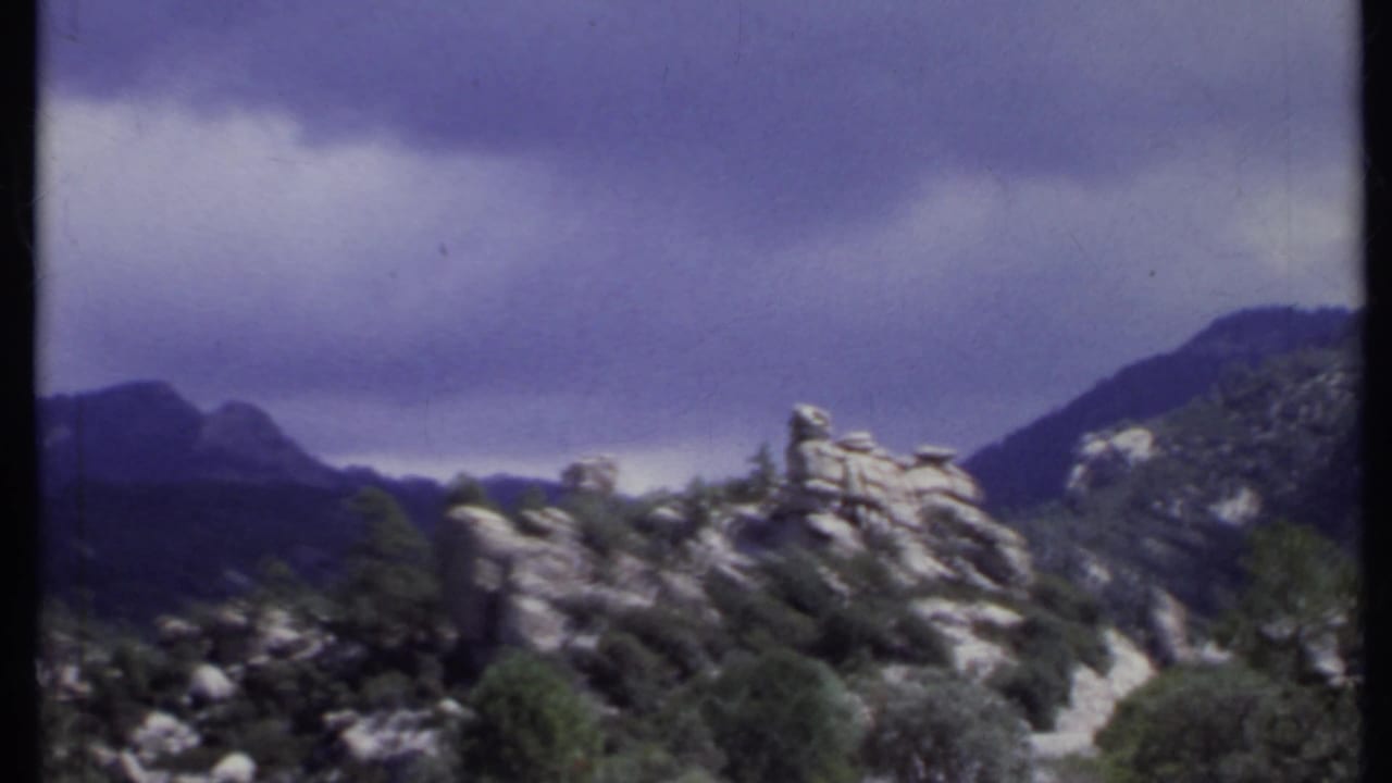 Rock outcrop, trees, and distant mountains under a dark cloud band.