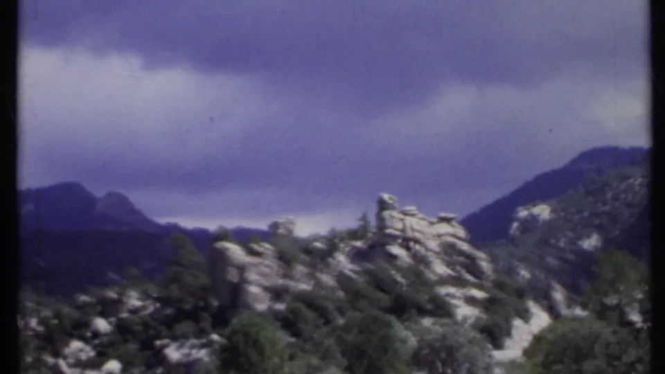 1971 California home-movie frame showing rugged rocks, tree cover, and distant mountain shapes beneath a dramatic overcast sky.