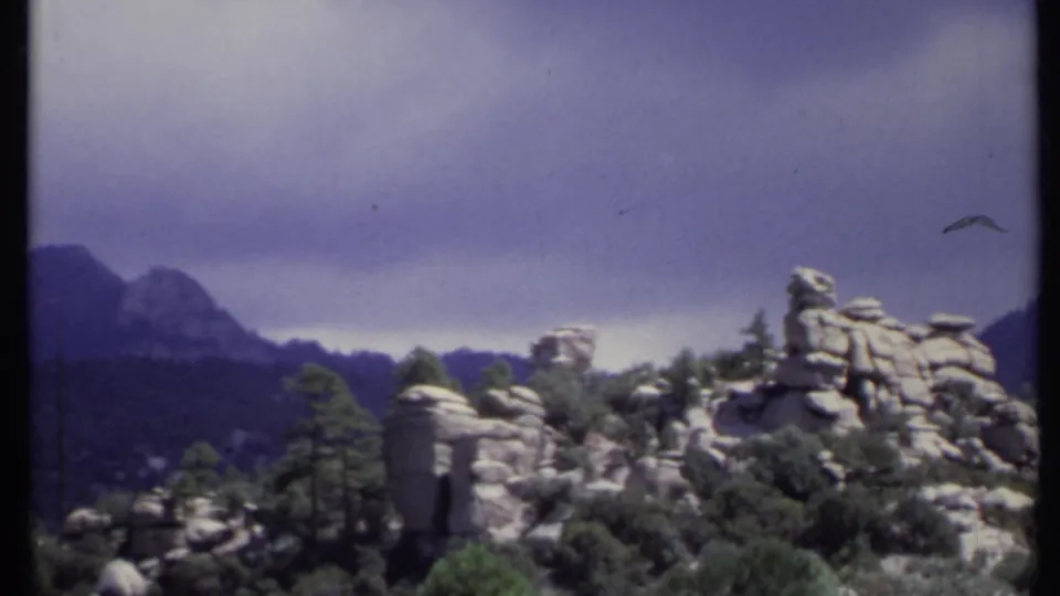 1971 California landscape frame with a prominent rocky outcrop, trees, and layered mountains beneath a purple-gray sky.