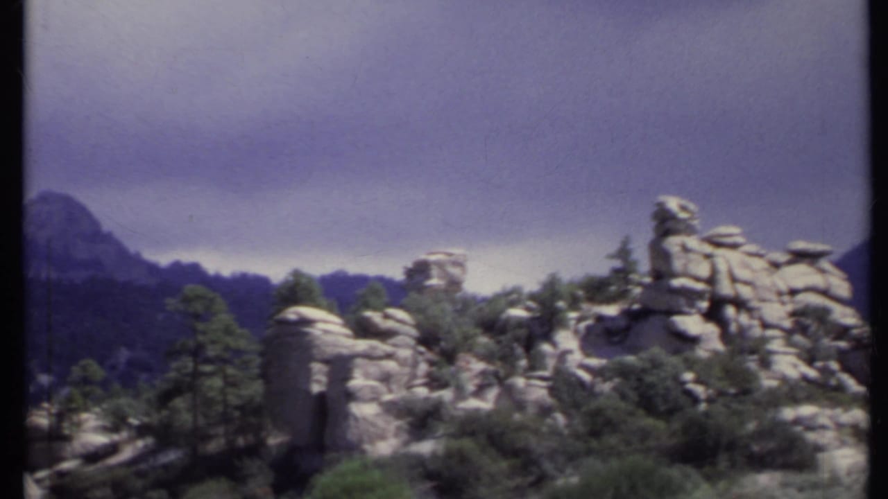 1971 California 8mm frame showing rugged rock formations, scattered pines, and distant hazy mountain ridges under a gray sky.