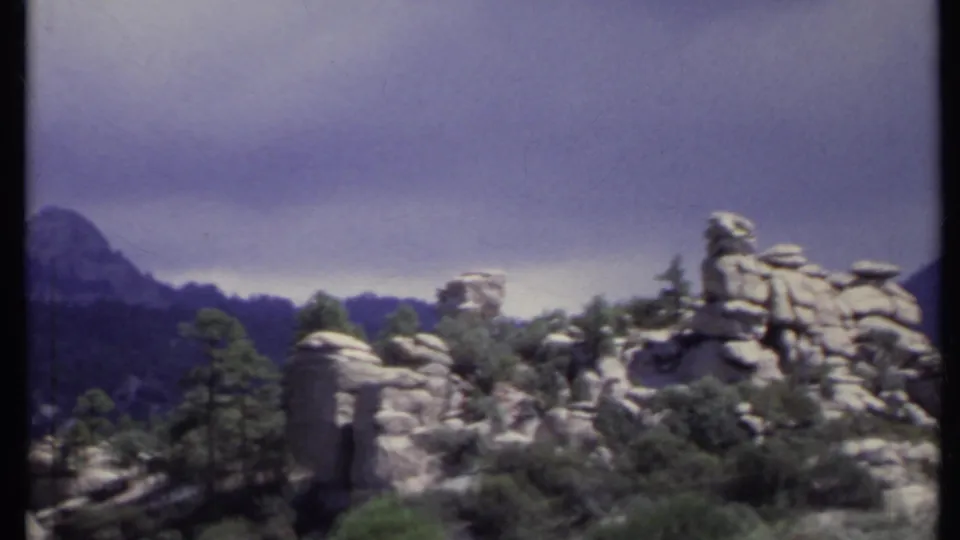 1971 California 8mm frame showing rugged rock formations, scattered pines, and distant hazy mountain ridges under a gray sky.