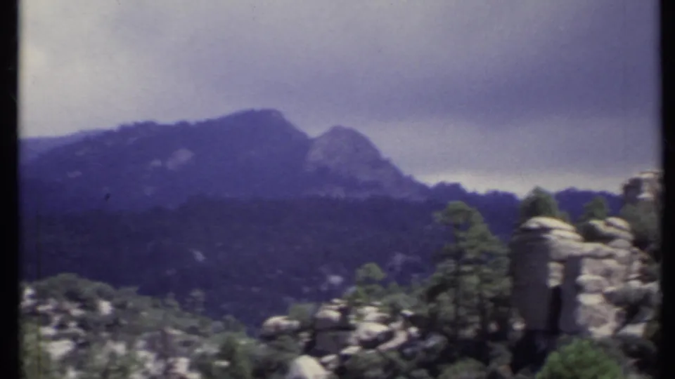 1971 California 8mm view of a dark mountain ridge rising beyond rocky, pine-dotted terrain under cloudy light.