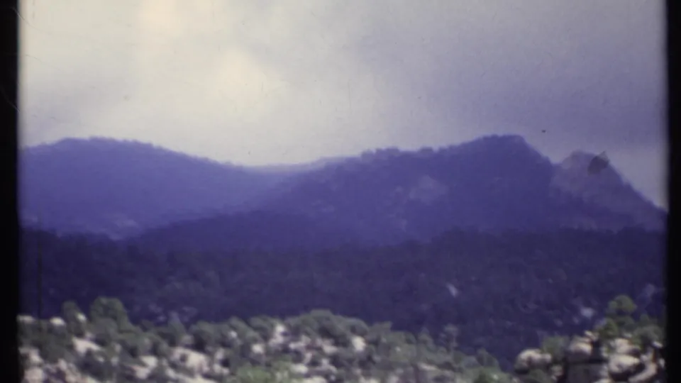 1971 California 8mm frame with soft blue-purple haze over layered mountain ridges, forested slopes, and foreground rocks.