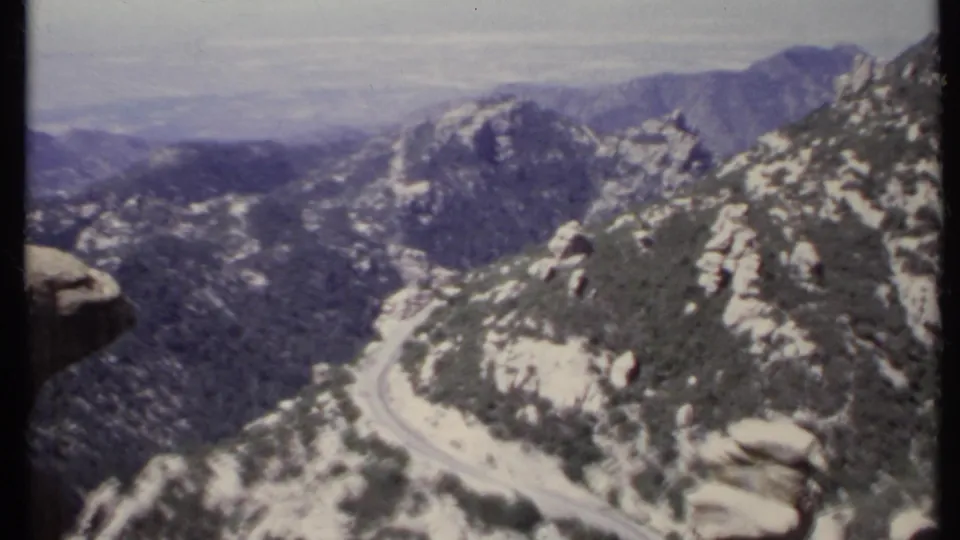 1971 California 8mm view of a pale winding mountain road below rocky slopes, pines, and distant hazy terrain.