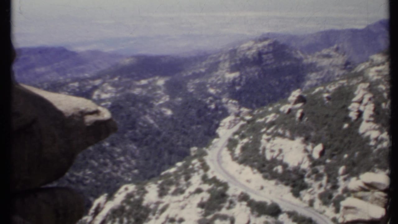 High mountain overlook with a winding road below, forested slopes, distant ridges, and a large rock at left.