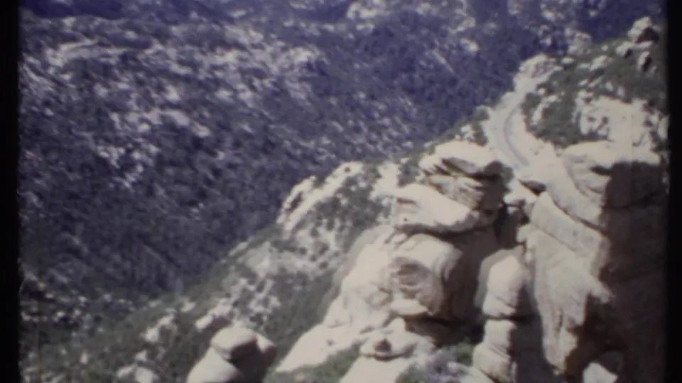 A 1971 California mountain view looking down past rounded rock formations toward a curving road and dense forested slopes.
