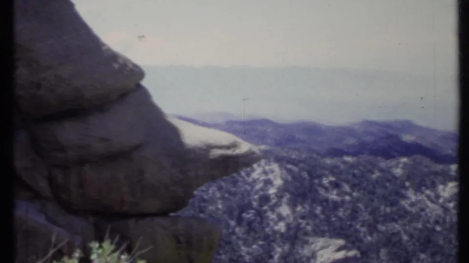 1971 California 8mm frame with a large foreground boulder overlooking forested mountain ridges and distant hazy terrain.