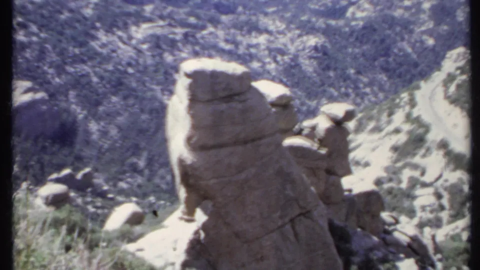1971 California home-movie frame of a tall rock outcrop rising over brush-covered sandy hills and a winding road.