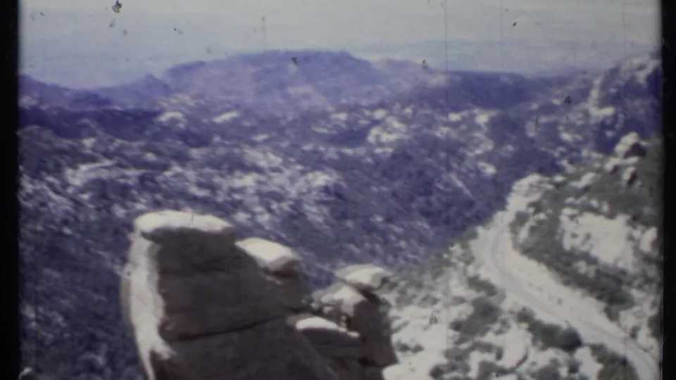 Wide 1971 California 8mm frame with eroded rock forms in the foreground, hazy hills, sparse brush, and a winding road.