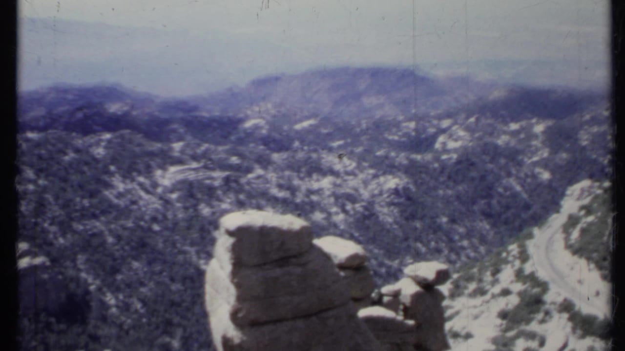 Rocky mountain landscape with a winding road below and boulders in the foreground.