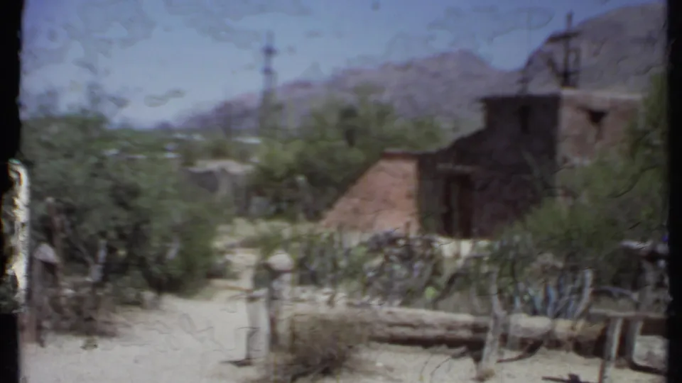 California 1971 home-movie frame showing an adobe-style structure, desert shrubs, fencing, and mountains in the background.