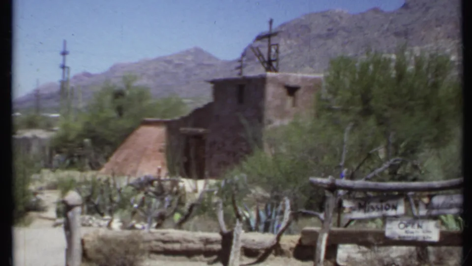 1971 California 8mm frame of mission-style architecture with cacti, a rustic fence, mountains, and a partially readable Mission Inn sign.