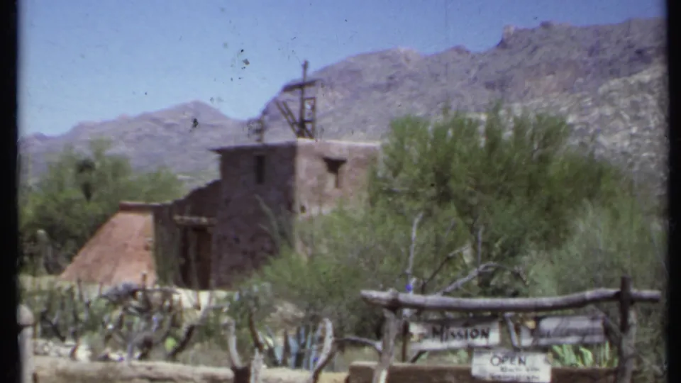 1971 California home-movie frame showing an adobe-style mission setting, desert landscaping, mountains, and partially readable roadside signage.