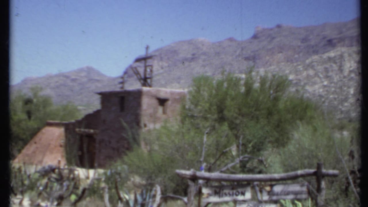 Adobe mission building with roof cross behind a wooden Mission Open sign and desert plants.