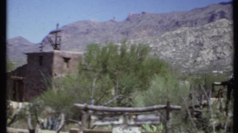 1971 California home-movie frame with the mission sign in front of an adobe structure, roof cross, desert vegetation, and mountains.