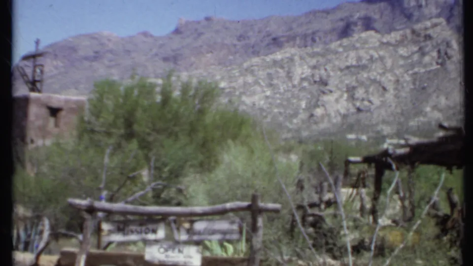 1971 California 8mm frame centered on the Mission Open sign, rustic fence, desert vegetation, and mountains, with the adobe building at left.