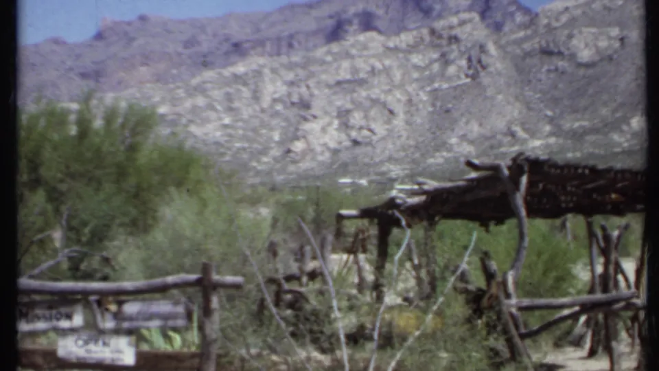 1971 California home-movie frame showing the mission grounds with rustic fencing, desert plants, a rough shade structure, and mountain backdrop.