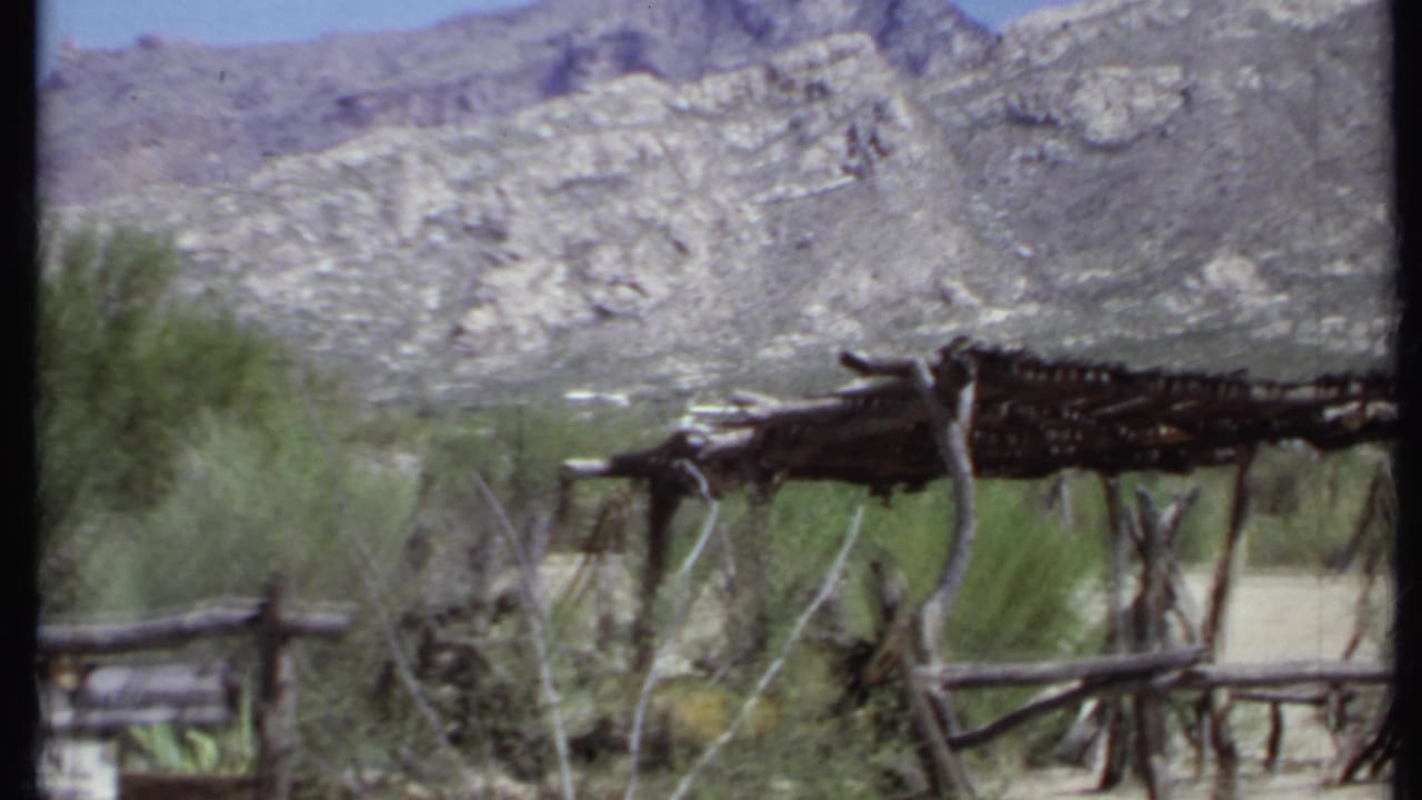 Rustic stick shelter and branch fence below rocky hills in a dry landscape.