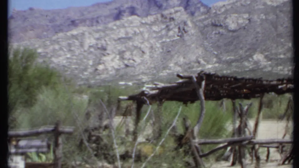 1971 California 8mm frame showing a rough stick shelter, branch fence, shrubs, dry ground, and rocky mountain slopes.