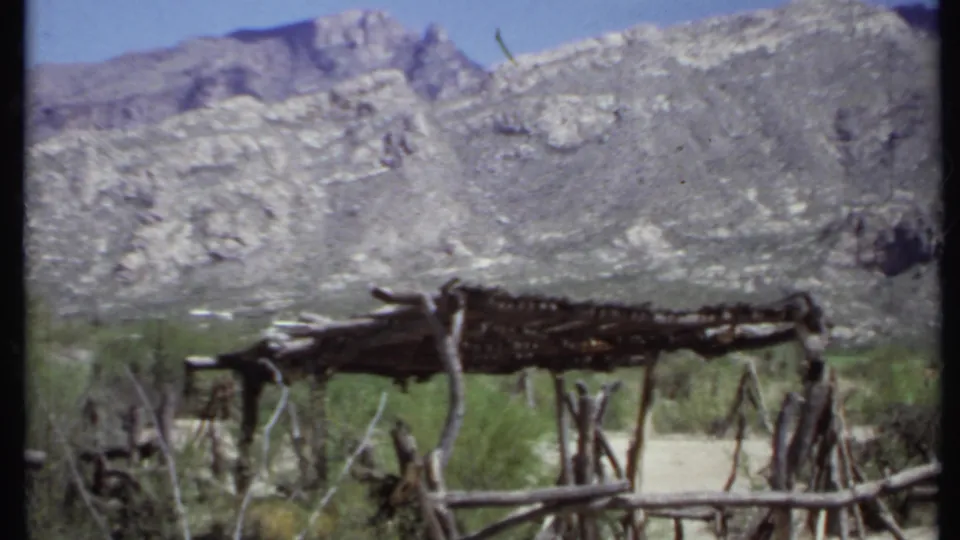 1971 California home-movie frame with a rustic branch hut set among shrubs and dry grass below rocky hills.