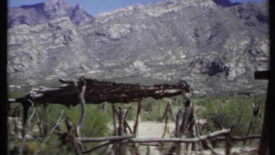 1971 California 8mm frame showing a rough wooden shelter and fence in front of a steep rocky desert hillside.