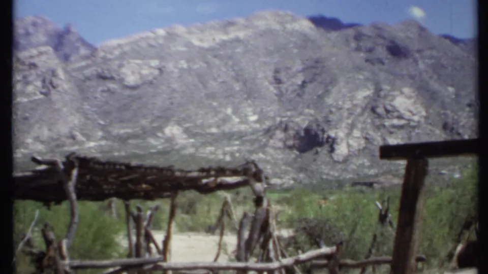 1971 California frame with a stick shelter, branch fence, shrubs, and rocky mountain terrain, partly obscured by film frame edges.