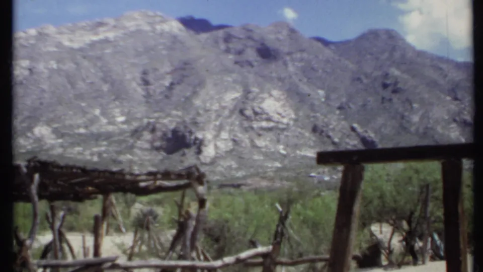 1971 California 8mm frame looking across a dirt roadside area with rustic fencing, desert shrubs, and a mountain range.