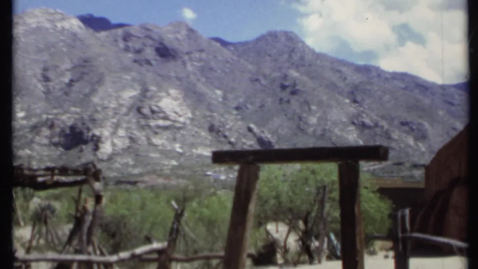 1971 California home-movie frame showing a timber fence, desert vegetation, rocky mountains, and part of a rustic structure at right.
