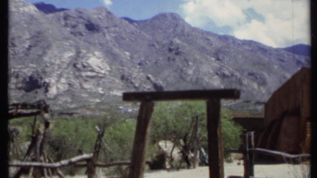 Adobe-style building edge, wooden gate, branch fence, shrubs, and rocky mountains.
