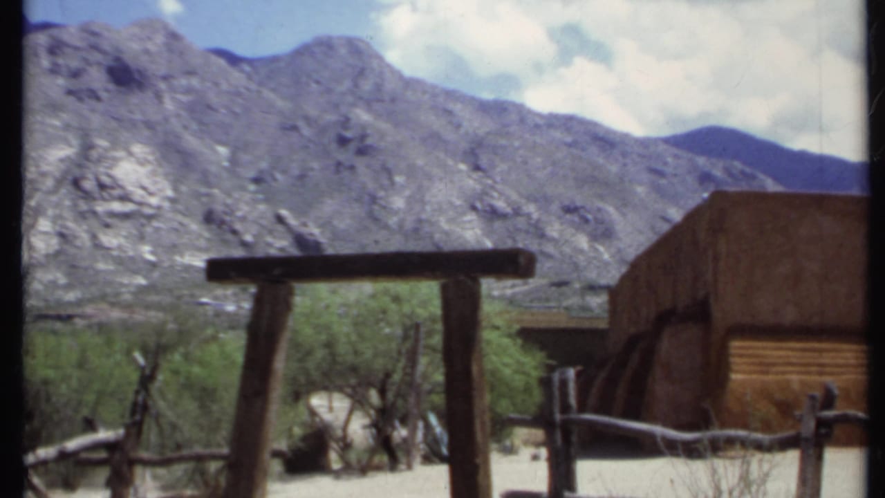 Wooden gate, adobe-style wall, branch fence, shrubs, and mountain ridge.