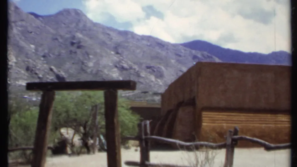 1971 home-movie frame of an adobe-style exterior, rustic fencing, sparse brush, and rugged California mountains.