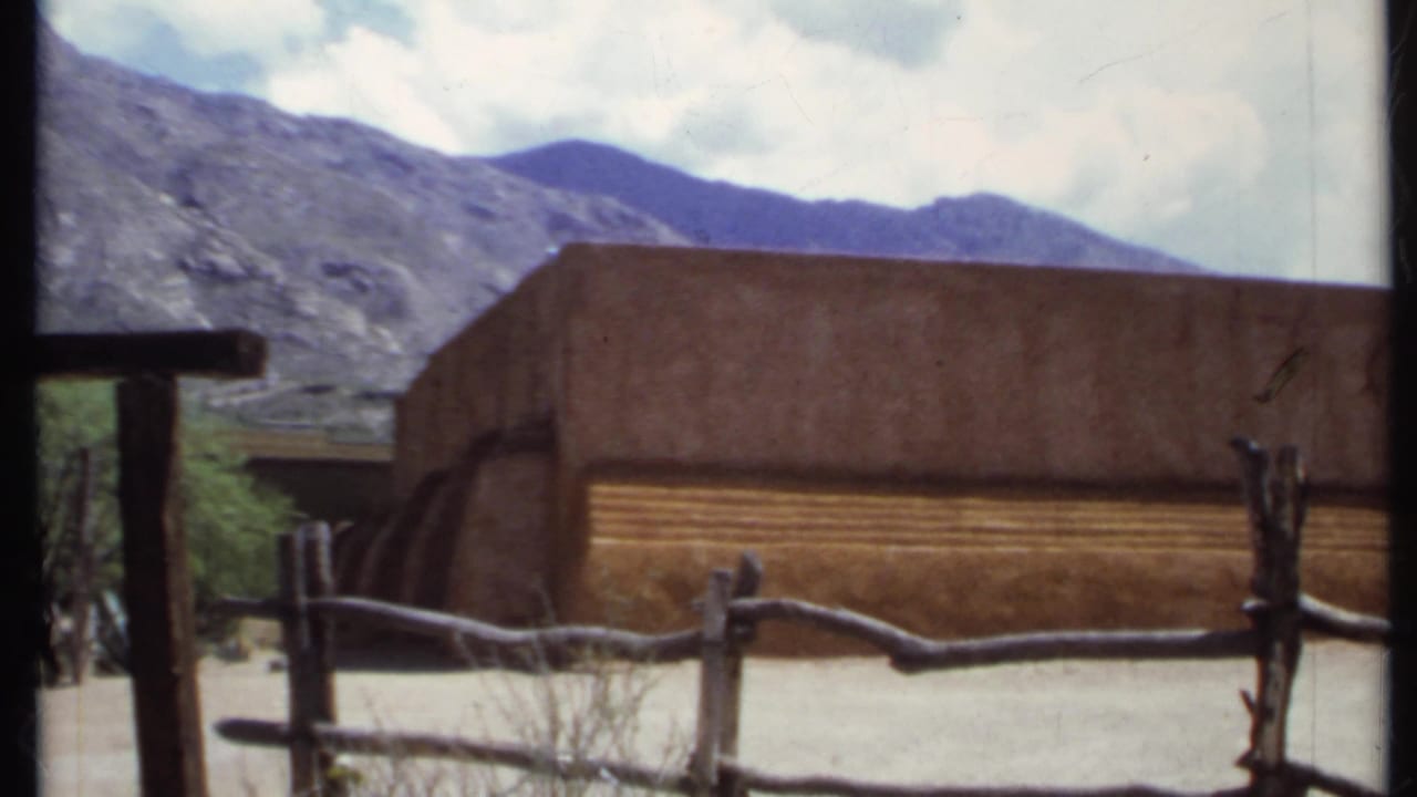Adobe-style building behind a rough wooden fence with mountains and cloudy sky beyond.