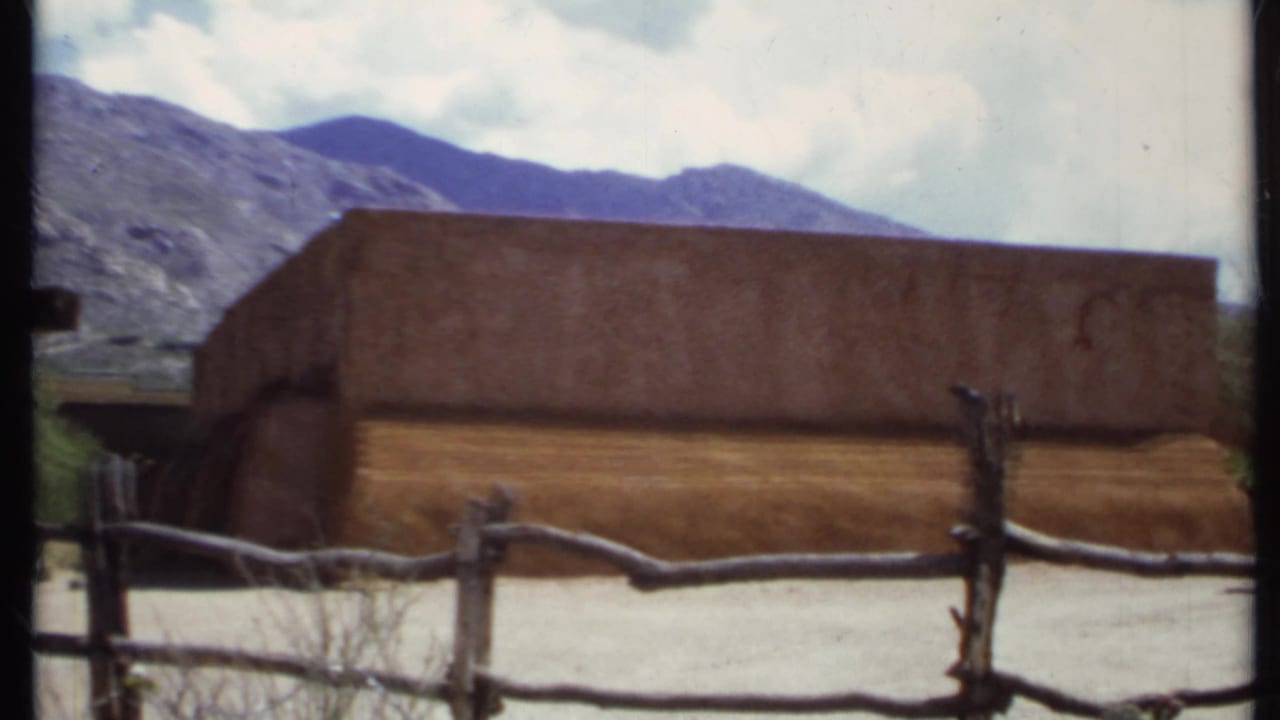 Similar view of the adobe-style building, fence, mountains, and cloudy sky.