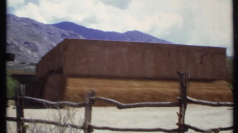 1971 California home-movie frame with an adobe-style building behind a rough fence and rugged mountains under a partly cloudy sky.