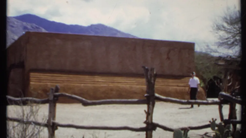 1971 California 8mm frame showing an adult man near the adobe-style building, with a rough fence, desert plants, and mountains visible.