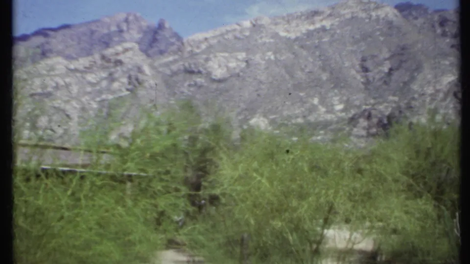 1971 California 8mm frame with a gray mountain ridge, blue sky, and windblown desert shrubs across the foreground.