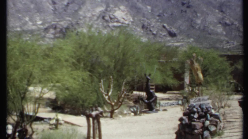 1971 California home-movie frame of a desert garden path with shrubs, cactus forms, rocks, fencing, and several dark outdoor sculptures.