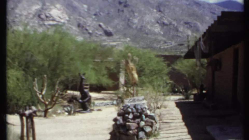 1971 California 8mm frame with desert shrubs, cactus, stone landscaping, a dark adobe-style structure edge, and a rocky mountain backdrop.