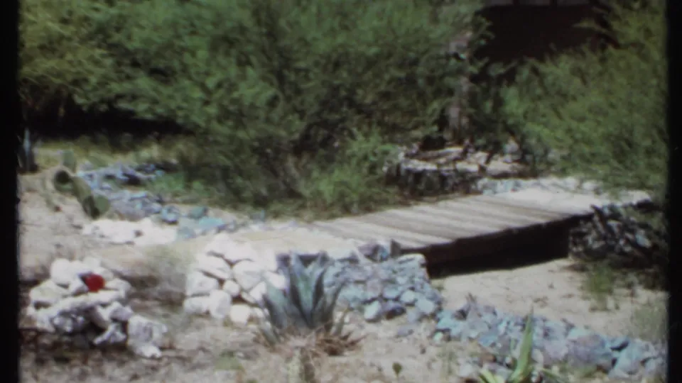 1971 California 8mm frame of a wooden plank bridge over a dry garden bed, with rocks, cactus, shrubs, and desert plants around it.