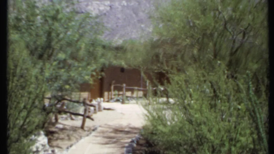 1971 California 8mm frame showing a sunlit dirt path, desert trees, rustic fencing, and an adobe-style structure in the distance.