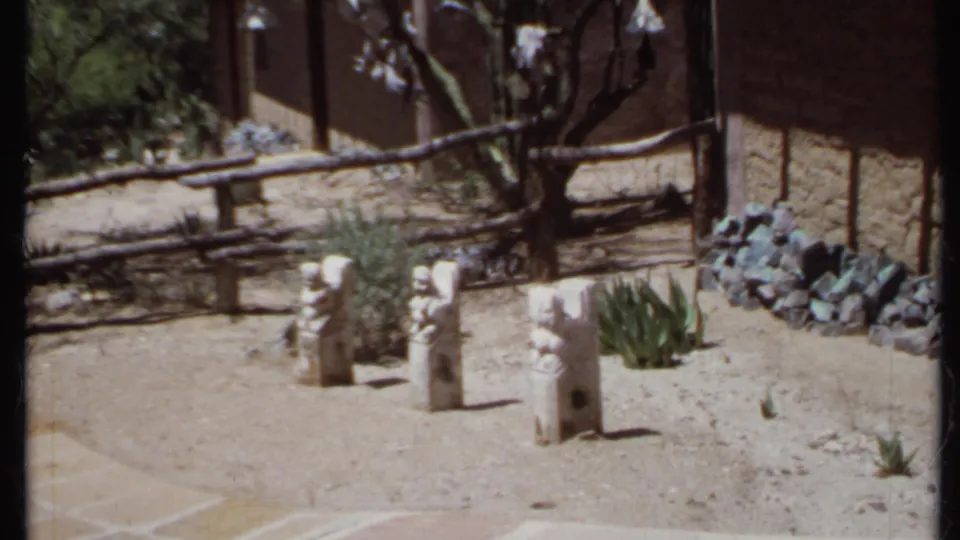1971 California 8mm frame looking down at stone yard sculptures in a dry landscaped area with cacti, rocks, rustic fence, and trees.