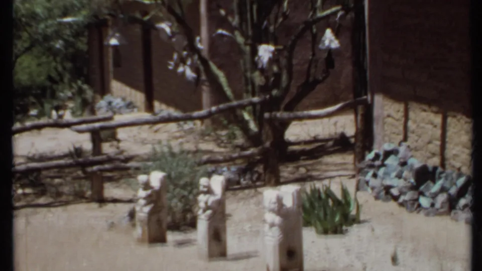 1971 California 8mm frame of a landscaped desert courtyard with three stone sculptures, cactus plants, rustic fencing, trees, and an adobe-style wall.
