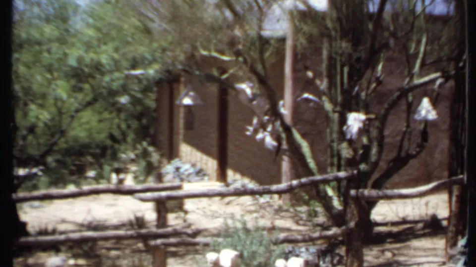 1971 California 8mm view of a dry meditation-like outdoor area with tree branches, rough wooden fencing, and an adobe-style building wall.