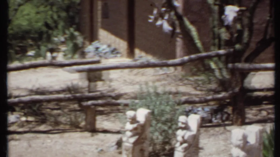 1971 California 8mm frame showing a sunlit dirt path, wooden rail fence, desert plantings, stone figures, and part of an adobe-style building.