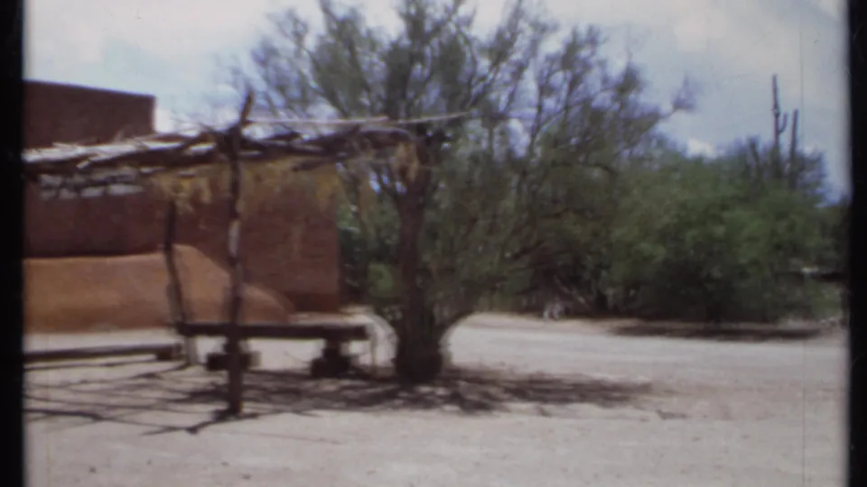1971 California 8mm view of an adobe-style wall, rustic wooden shade frame, sandy ground, and desert vegetation.
