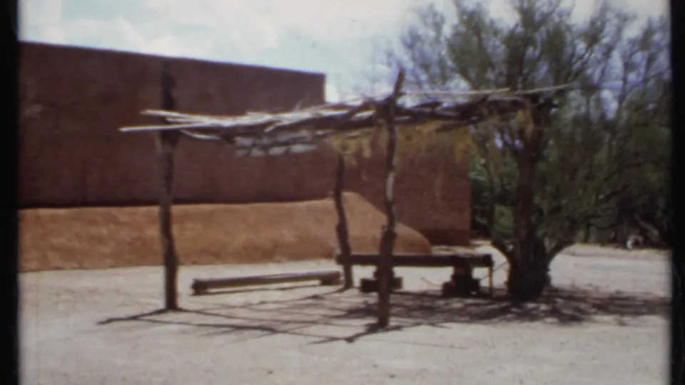 1971 California frame showing a pole-built shade structure, adobe-style walls, bare ground, and sparse desert trees.