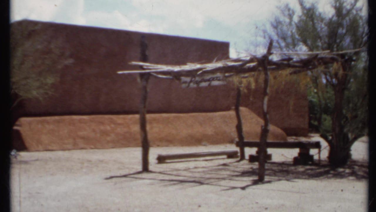 Adobe exterior with wooden shade arbor, bench, desert plants, and pale ground.
